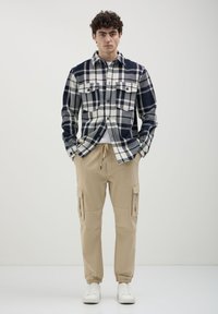 Camicia a scacchi blu e bianca con due tasche sul petto, pantaloni cargo beige con polsini elasticizzati, abbinati a sneakers bianche. Design semplice e funzionale.