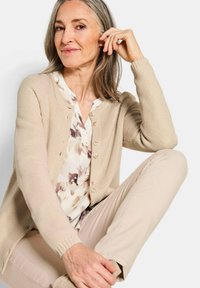 Cardigan beige en tricot avec un tissu texturé, col rond et fermeture par boutons. Associé à un blouse à motifs et un pantalon beige clair.