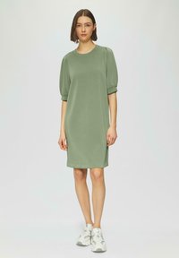 Grünes Kurzarmkleid mit lockerer Passform, Rundhalsausschnitt und gerippten Akzenten. Getragen mit weißen Sporttrainern.