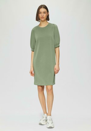 Groene jurk met korte mouwen en een relaxte pasvorm, met een ronde halslijn en geribbelde accenten. Draagt witte sport sneakers.