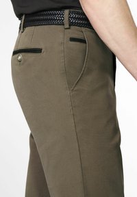 Close-up van een persoon die bruine broek draagt met een zwart gevlochten riem en zwarte randen op de achterzakken, zijaanzicht van taille tot bovenbeen.