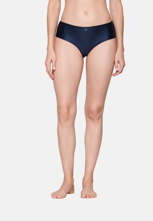 Woman wearing navy blue ribbed mid-rise brief underwear, standing barefoot against a plain light background.