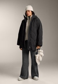 Person trägt eine schwarze gesteppte Jacke, graue Strickhose, weiße Mütze und hält cremefarbene, flauschige Hausschuhe vor schlichtem Hintergrund.