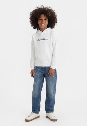 Uśmiechnięte dziecko o kręconych włosach, ubrane w białą bluzę z kapturem Calvin Klein, niebieskie jeansy i białe tenisówki, stojące na jednolitym tle.