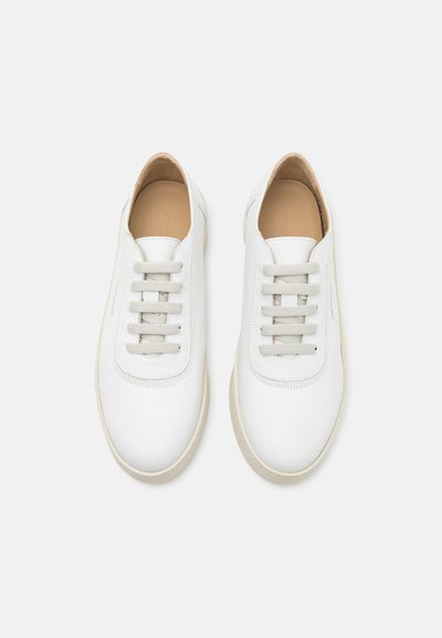 Zapatillas de cuero blancas con una textura suave, punta redondeada y cordones grises sutiles. Presentan una suela de goma beige clara y un branding mínimo.