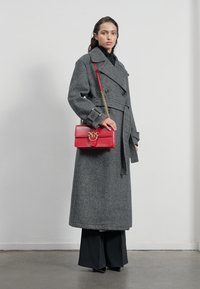 Sivý vlnený trenčkot s dvojradovým dizajnom, širokými klopami, viazaným pásom, v kombinácii s malou červenou koženou kabelkou so zlatými kovovými doplnkami.