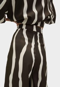 Femme portant un pantalon taille haute et un haut assorti avec des rayures verticales ondulées en noir et blanc, posant les mains sur les hanches.