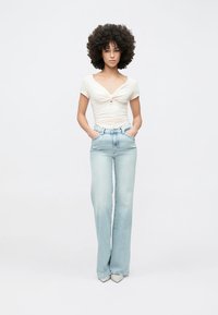 Hvit, tettsittende topp med knutedetalj foran, kombinert med lyseblå vide jeans. Jeansene har høyt liv og klassiske fem lommer.