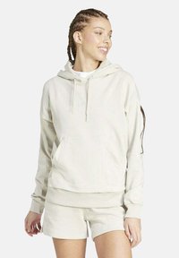 Giovane donna con capelli intrecciati che indossa un felpa beige e pantaloni corti abbinati, sorridente e con lo sguardo rivolto di lato su uno sfondo bianco.