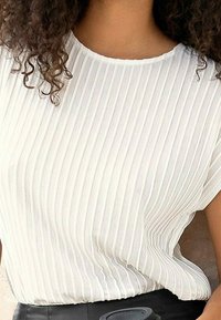 Kvinna iförd en vit ribbad t-shirt med kort ärm och lockigt hår synligt på axlarna.