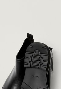 Paire de bottines en cuir noir, l’une présentant une tige lisse, l’autre montrant une semelle en caoutchouc avec un profilé à motif et une languette en tissu.