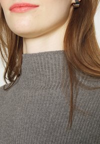 Pull à col roulé gris côtelé avec une coupe ajustée, un tissu texturé et un col haut. Les cheveux tombent partiellement sur l'épaule.