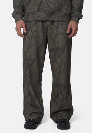 Uomo che indossa pantaloni larghi grigio scuro e felpa abbinata con un sottile motivo astratto, scarpe da ginnastica nere visibili sotto i pantaloni.