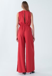 Rød ærmeløs buksedragt med tilpasset talje og vid ben. Har en udskæring midt på ryggen og en glat stoftekstur.