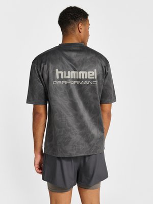 Mand iført mørkegrå "hummel PERFORMANCE" kortærmet skjorte og lagdelte mørkegrå træningsshorts, vendt væk mod en ensfarvet baggrund.