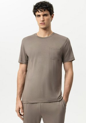 Mann trägt ein einfarbiges taupefarbenes Kurzarmhemd mit Brusttasche und passende Hose, steht vor weißem Hintergrund.