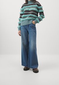 Turkoosi neulepaita, jossa on hapsukoristeet ja raitakuvio, yhdistetty leveälahkeisiin sinisiin farkkuihin ja mustiin korollisiin kenkiin.