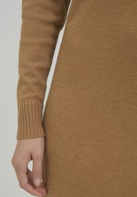 Pulover tricotat de bronz, cu mâneci lungi, manșete ribbate și o textură moale. Prim-planul arată mâna odihnindu-se pe material.