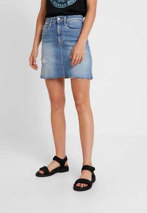 Person iført en lyseblå denim miniskørt med frynset kant og sorte sandaler, stående mod en ensartet lys baggrund.