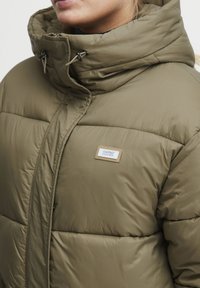 Doudoune vert olive avec un col haut, présentant un motif cousu et un écusson de logo sur la poitrine. Cordons de capuche ajustables.