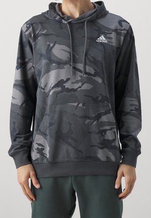 Homme portant un sweat à capuche Adidas gris foncé à motif camouflage avec poche avant et capuche à cordon, associé à un pantalon vert foncé.