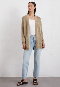 Beige gebreide cardigan met open voorkant, ribgebreide manchetten en zoom, gedragen over een witte top en lichtblauwe rechtlopende spijkerbroek, gecombineerd met zwarte sandalen.
