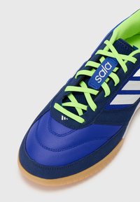 Zapatilla deportiva azul con cordones verdes, detalles de malla y una suela de goma texturizada. Presenta tres rayas blancas a un lado y una etiqueta de logo.