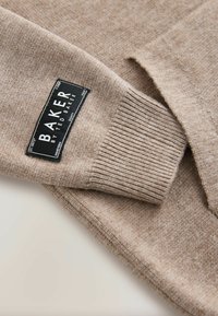 Brązowy sweter dzianinowy z prążkowanymi mankietami. Posiada prostokątną czarną metkę z białym tekstem informującym o marce i instrukcjach dotyczących pielęgnacji.