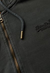 Felpa zip grigia scura con zip dorata, tessuto testurizzato e un piccolo logo ricamato. Presenta coulisse grigie coordinate e dettagli metallici.
