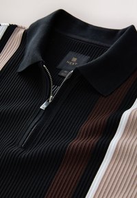 Polo en maille côtelée noire avec fermeture éclair, présentant des rayures verticales marron, blanches et beige sur les manches, et une petite étiquette noire à l'intérieur du col.