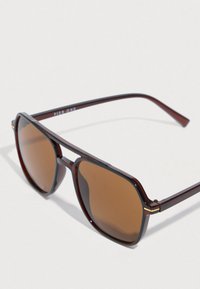 Smeđe sunčane naočale s tamnosmeđim okvirom u uzorku kornjačevine, geometrijskog oblika i tankim ručkama s zlatnim detaljima.