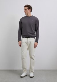 Un homme se tient en regardant vers la gauche, portant un sweatshirt gris foncé, un pantalon écru, une ceinture noire et des baskets blanches devant un fond gris uni.