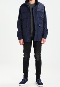 Chaqueta ligera de color azul marino con cuatro bolsillos frontales, botones a presión y un cuello alto. Lleva puesta sobre un suéter gris y combinada con jeans negros y zapatillas.