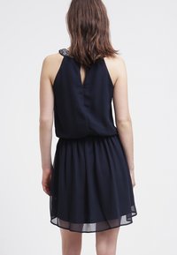 Marinblå klänning med ärmlös design, rynkad midja och nyckelhål bak. Utsmyckad med en dekorativ detalj på axeln.