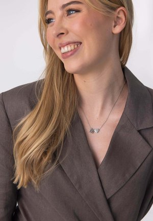 Lächelnde blonde Frau mit langen Haaren, die einen braunen Blazer und eine silberne Halskette mit einem "M"- und Herzanhänger trägt.