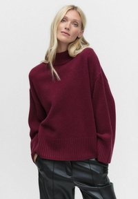 Burgundy tröja med hög hals, oversize passform, ribbade muddarna och f�ållen, i kombination med svarta läderbyxor, mot en vit bakgrund.