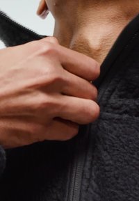 Ruka hvata patentni zatvarač na teksturiranoj crnoj flis jakni blizu golog vrata.