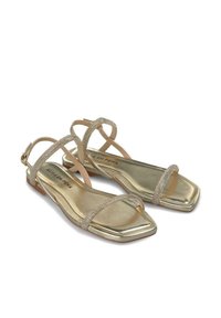 Złote płaskie sandały o błyszczącej metalicznej wykończeniu. Posiadają krzyżujące się paski z ozdobami w postaci cyrkonii oraz kwadratowy nosek.