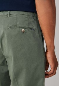 Pantalon vert kaki avec une texture lisse, doté d'une poche arrière avec fermeture à bouton et d'une petite étiquette bleue.