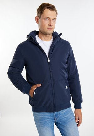 Man in een marineblauwe rits-hoodie en lichtblauwe spijkerbroek, staande met één hand in de zak tegen een witte achtergrond.