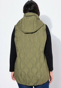 Gilet trapuntato verde oliva con cappuccio, caratterizzato da un motivo a onde testurizzato, orlo basso e una maglietta nera a maniche lunghe sotto.