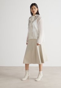 Weißes transparentes Blusenshirt mit Rüschensaum, kombiniert mit einem beigen Midirock und weißen Absatzstiefeletten. Glatter Stoff, fließende Silhouette.