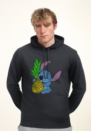 Jonge man met een zwarte hoodie waarop een afbeelding staat van een blauwe cartoonfiguur die een grote ananas omhelst.