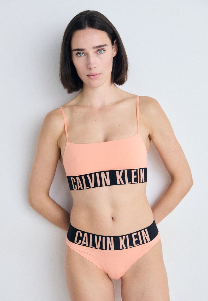 Calvin Klein bikini-set in perzik met dunne bandjes, een vierkante halslijn en een brede zwarte tailleband met de merknaam in roze letters.
