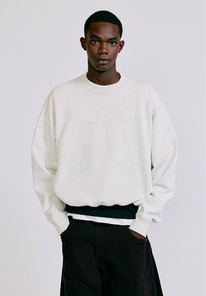 Junger Mann trägt einen hellgrauen übergroßen Pullover und schwarze Hose mit den Händen in den Taschen, blickt nach vorne auf einem schlichten Hintergrund.
