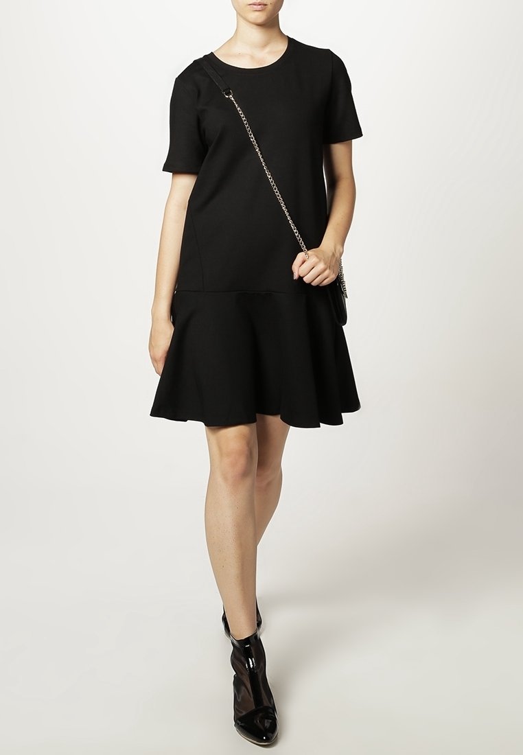 Robe noire à manches courtes avec un bas évasé, en tissu lisse. Associée à des bottes noires brillantes, arrivant aux chevilles.