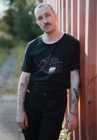 Camiseta negra de manga corta con un gráfico geométrico de montaña en blanco y rosa. Combinada con pantalones negros, que presentan herrajes plateados, y tatuajes visibles.