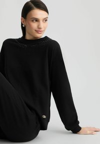 Zwarte sweatshirt met een textuur van parelontwerpen rond de halslijn, lange mouwen en een klein gouden logo-accent nabij de zoom.