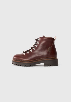 Stivaletto in pelle marrone con suola nera robusta, occhielli in metallo, lacci marroni e neri, e linguetta sul retro su sfondo bianco.