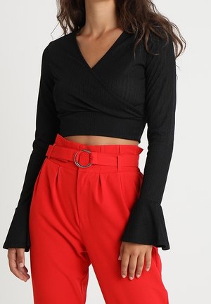 Nainen, jolla on musta pitkähihaisen lyhyen topin liehuvilla kalvosimilla ja korkeavyötäröiset punaiset housut, joissa on vyö ja hopeinen solki.